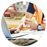 Construction worker holding coffee checking laptop and blueprints on site.