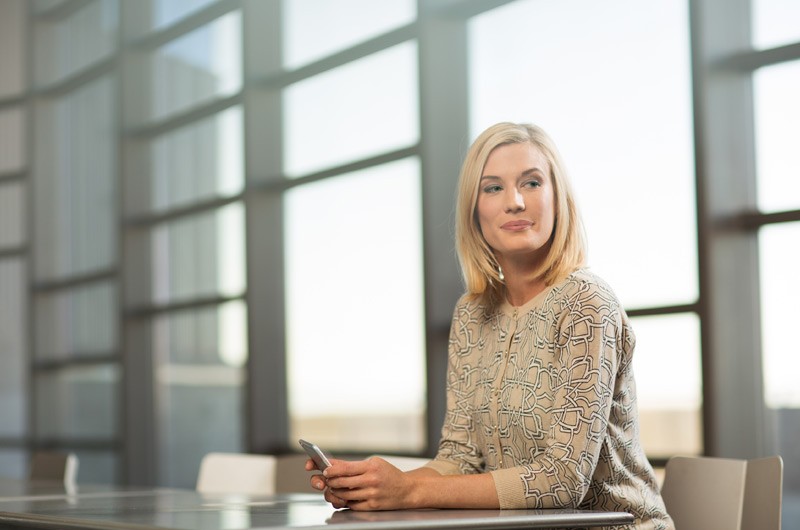 Attractive woman sitting in room with large windows holding smart phone.