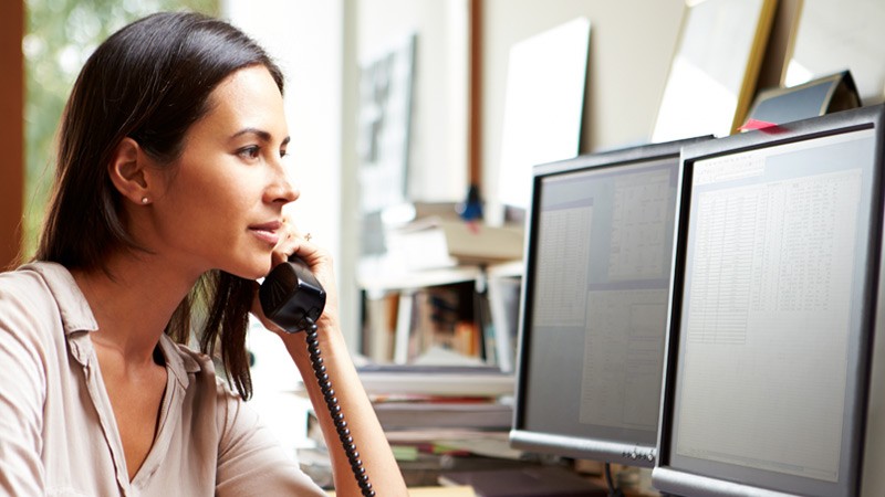 woman-on-wired-phone-at-desk-two-monitor-displays