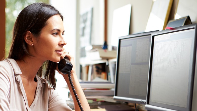 woman-on-wired-phone-at-desk-two-monitor-displays woman-on-wired-phone-at-desk-two-monitor-displays
