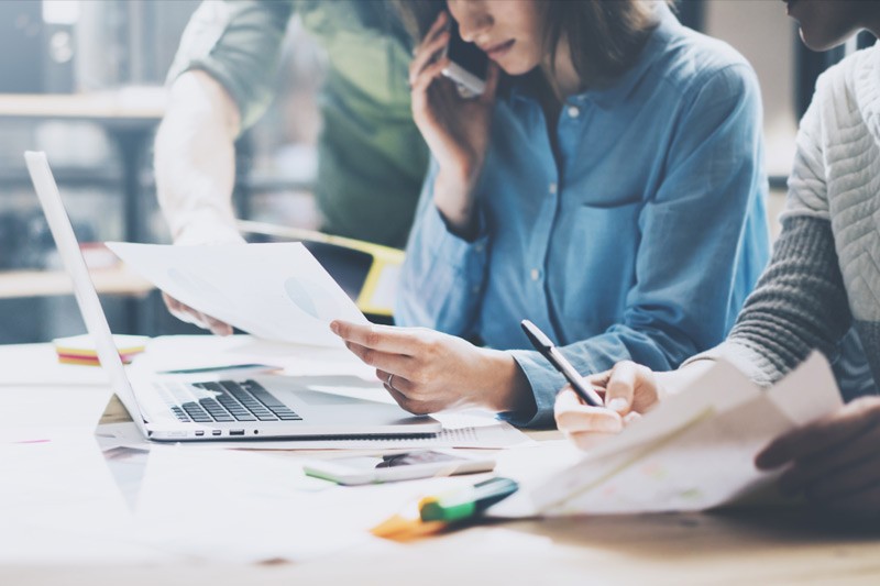 Young professional woman sitting at laptop looking over a printout she's holding in her hand.