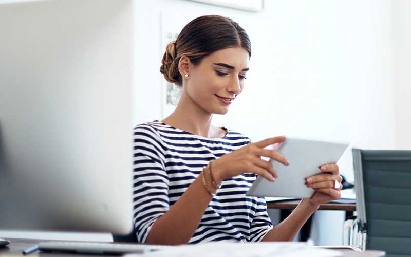 Femme d’affaires souriante utilisant une tablette près d’un ordinateur, bénéficiant de l’intégration transparente des données.