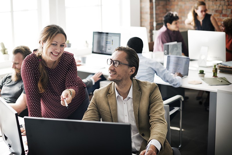 Business manager speaking with employee at desktop computer while team works behind them