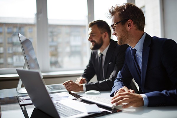 Two smiling businessmen look over desktop computers